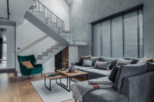 Ein Wohnzimmer mit einem grauen Ecksofa, einem grünen Samtsessel, zwei ineinander verschachtelten Couchtischen und einer Treppe, unter der ein Weinregal eingebaut ist.
