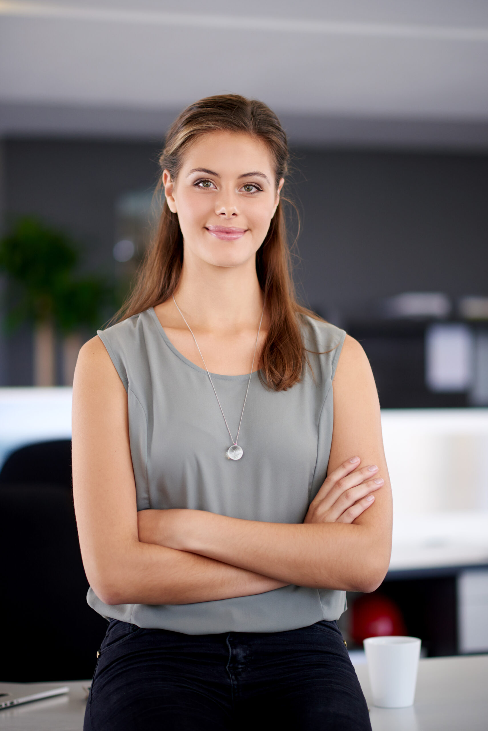 Eine junge Frau in einem grauen, ärmellosen Oberteil, die mit verschränkten Armen in einem Büro steht.