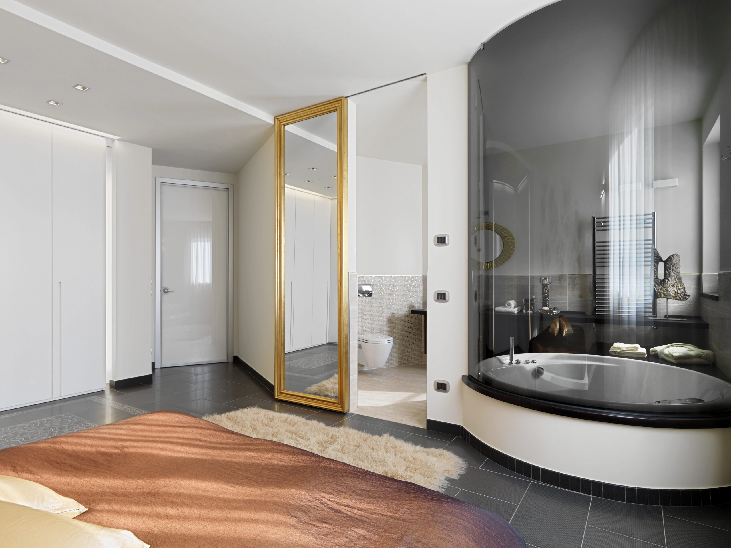 Schlafzimmer mit brauner Tagesdecke, großem goldgerahmtem Spiegel und angrenzender verglaster Badewanne.