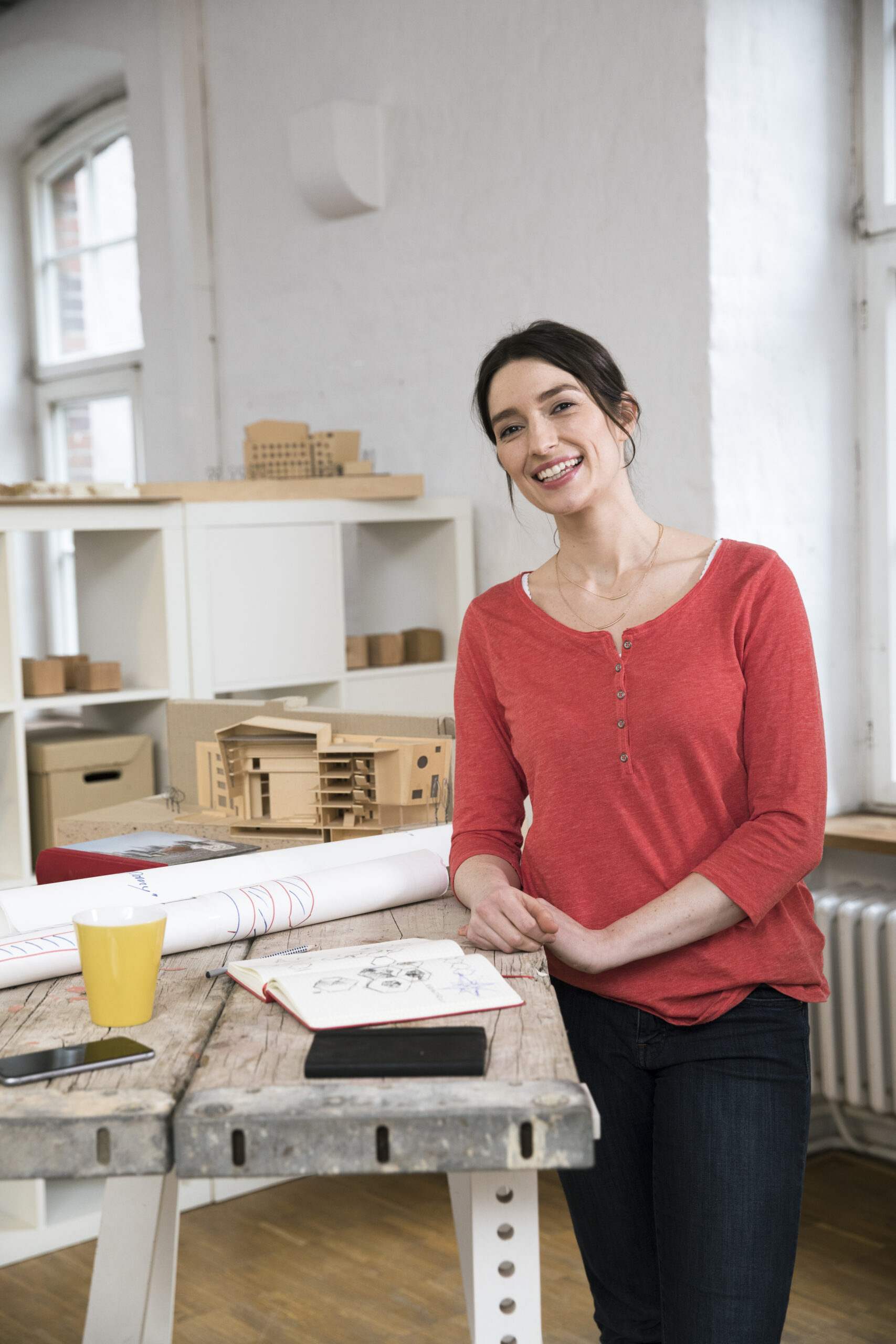 Eine lächelnde Frau in einem roten Hemd steht an einem Zeichentisch, auf dem Architekturmodelle und ein Skizzenbuch liegen.