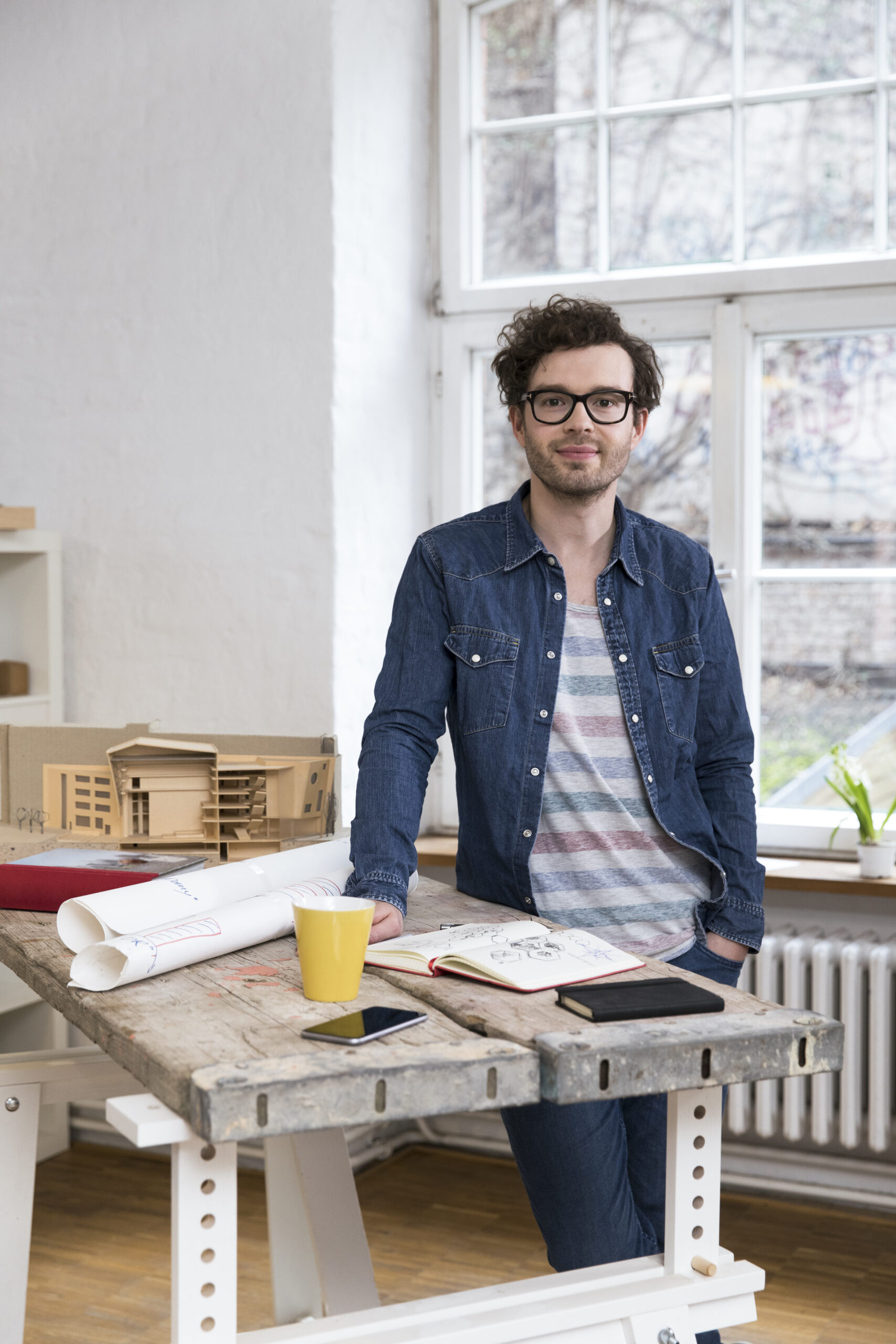 Ein Mann in einem Jeanshemd steht an einer hölzernen Werkbank, auf der ein Architekturmodell, ein Skizzenbuch und eine gelbe Tasse liegen.
